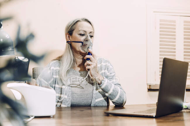 Старшая женщина принимает онлайн косультации во время вдыхания через распылитель на дому — стоковое фото