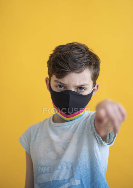 Gros plan du garçon portant un masque multicolore pointant tout en se tenant debout sur fond jaune — Photo de stock