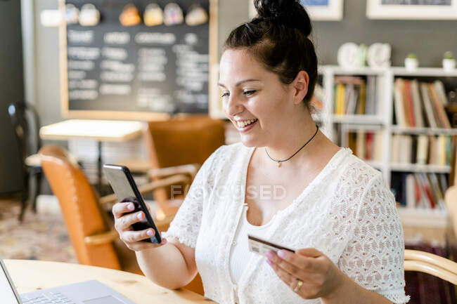 Close-up de sorrir jovem segurando cartão de crédito usando telefone inteligente no café — Fotografia de Stock