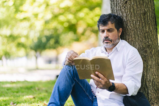 Зрелый мужчина читает книгу, опираясь на ствол дерева в общественном парке — стоковое фото
