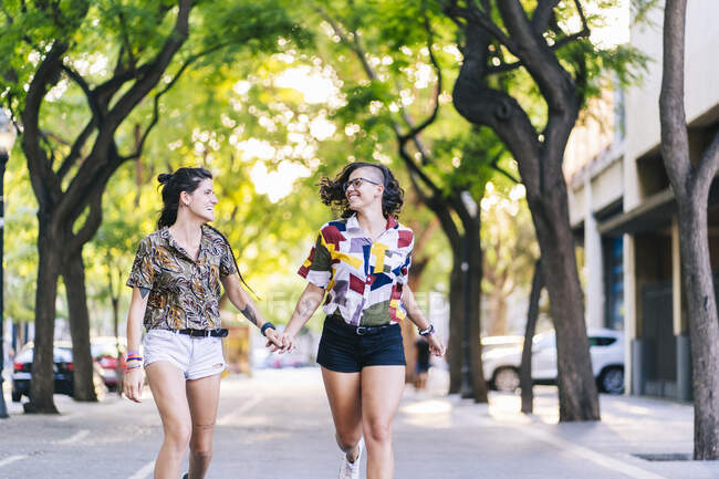 Felice coppia lesbica che si tiene per mano e corre sul sentiero in città durante la giornata di sole — Foto stock
