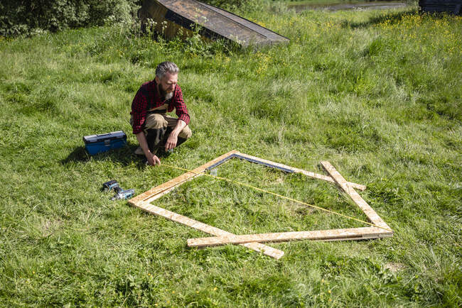 Carpenter crouching while measuring planks for playhouse on grass — Foto stock