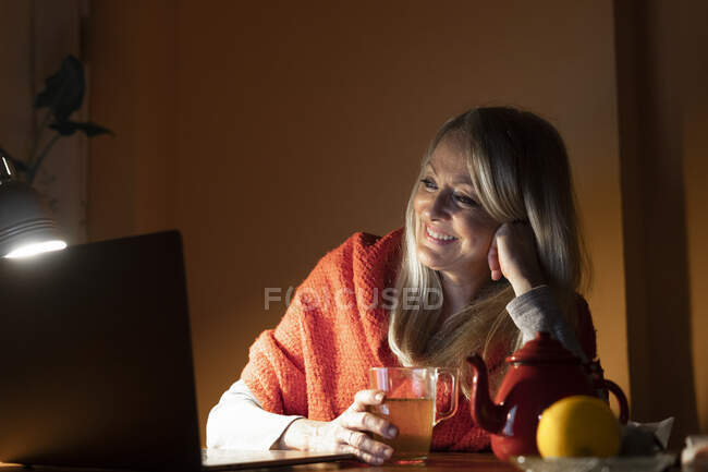 Smiling woman drinking tea while listening to video call on laptop at home — Stock Photo