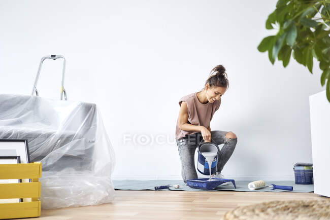 Sorrindo jovem mulher derramando tinta na bandeja de pintura enquanto se ajoelha em casa — Fotografia de Stock