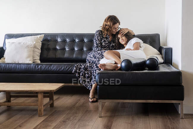 Madre abrazando a la hija mientras descansa en el sofá en casa - foto de stock