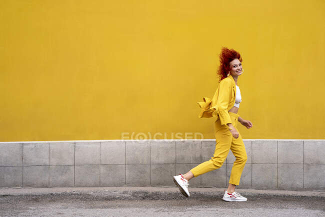 Energetic young woman in yellow suit running and jumping in front of yellow wall — Foto stock