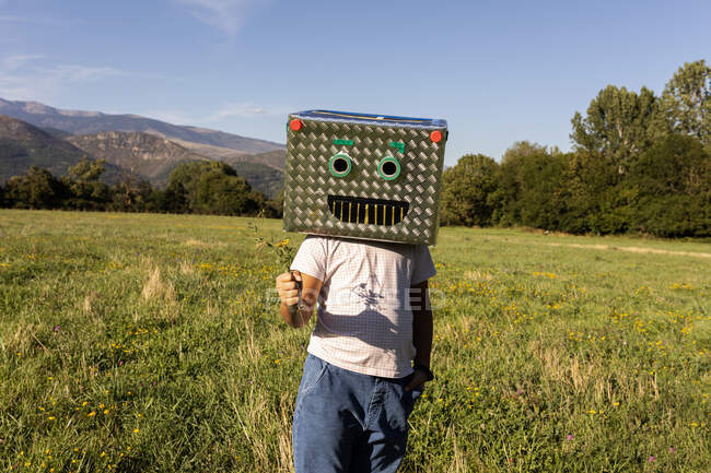 Мальчик с улыбающейся картонной коробкой на лице, держащий траву, стоя на лугу — стоковое фото