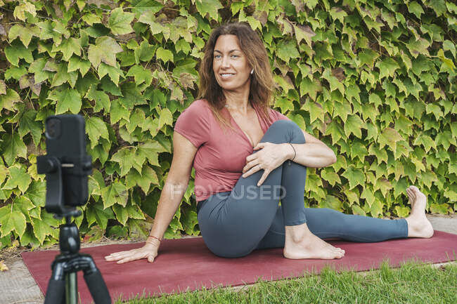 Frau streckt Körper während Online-Yoga-Kurs per Handy auf Stativ gegen Efeuwand — Stockfoto