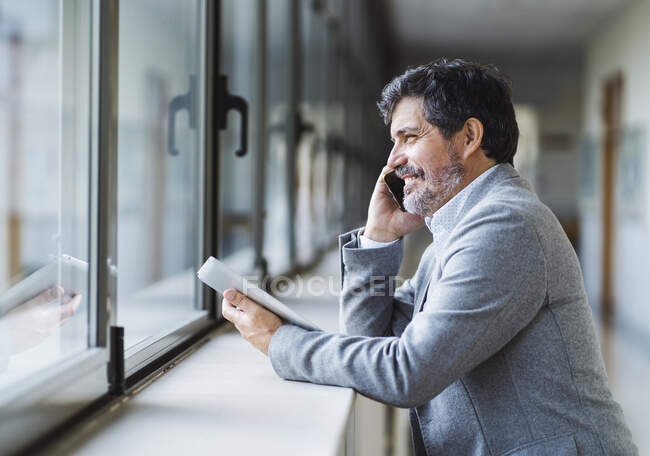 Lächelnder Professor telefoniert, während er durch das Fenster im Flur der Universität blickt — Stockfoto