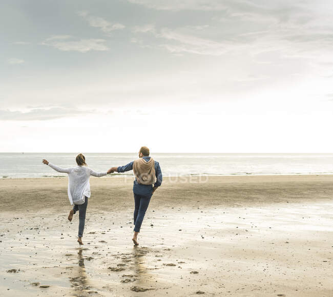 Весела зріла пара тримається за руки, стрибаючи на пляж проти хмарного неба. — стокове фото