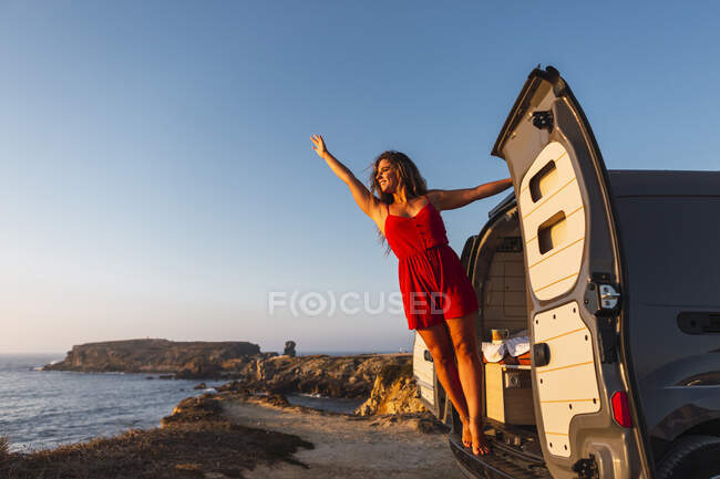 Donna sorridente con mano alzata in piedi mentre appeso sulla porta camper in spiaggia — Foto stock