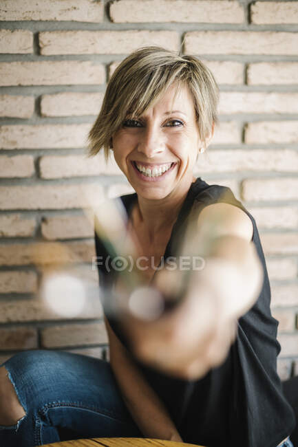 Mujer alegre mostrando pincel mientras se sienta contra la pared de ladrillo en casa - foto de stock