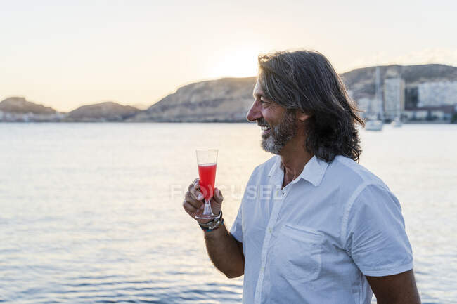 Sorridente uomo maturo che tiene bere guardando lontano mentre in piedi contro il mare durante il tramonto — Foto stock
