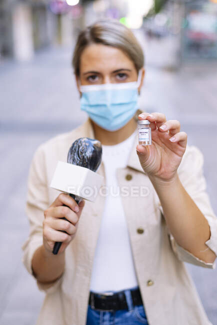 Female journalist wearing mask showing vaccine while standing on street in city — Stock Photo