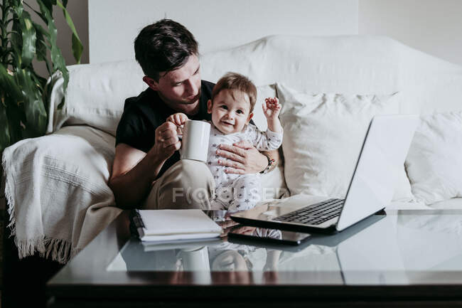 Pai segurando xícara de café com o filho sentado no sofá em casa — Fotografia de Stock