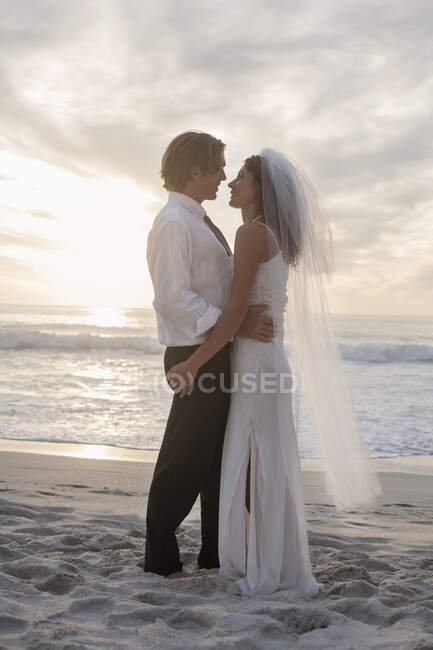 Couple nouvellement marié se regardant tout en se tenant sur le sable pendant le coucher du soleil — Photo de stock