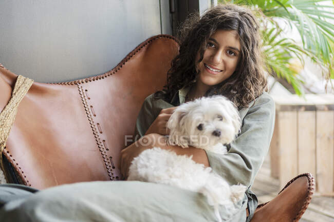 Ragazza sorridente che tiene il cucciolo mentre si siede sulla sedia a casa — Foto stock