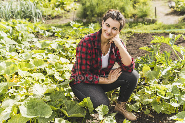 Молодая женщина с рукой на подбородке на коленях среди овощей, растущих в саду — стоковое фото