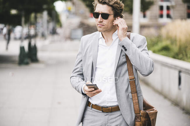 Businessman connecting in-ear headphones with mobile phone while standing at sidewalk in city - foto de stock