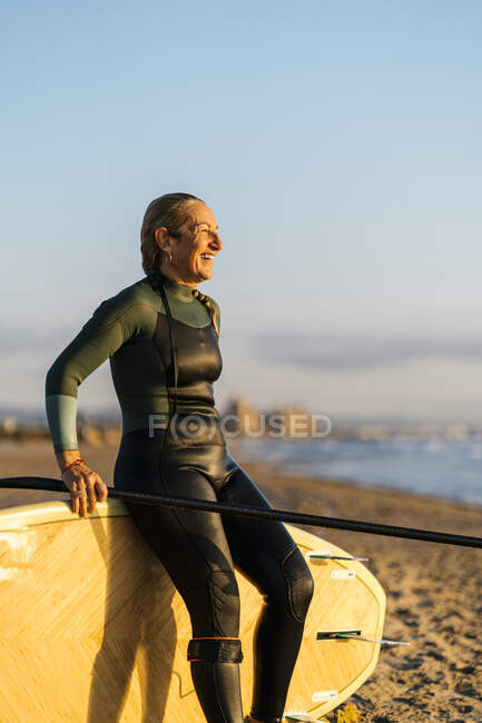 Frau lacht und schaut weg, während sie sich im Morgengrauen am Strand auf ein Paddelbrett lehnt — Stockfoto
