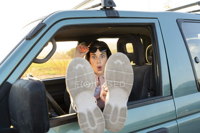 Удивлённая молодая женщина, сидящая с поднятыми ногами на окне машины во время заката — стоковое фото