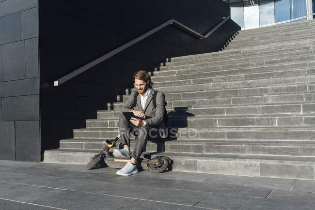 Businessman using digital tablet while sitting with electric push scooter on steps in financial district - foto de stock