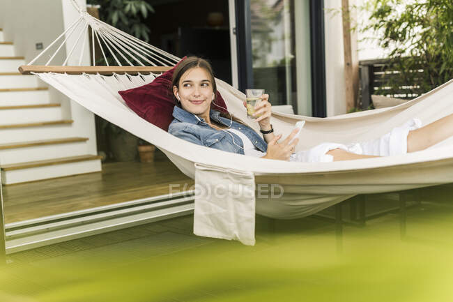 Beautiful woman holding mobile phone and drink looking away while resting on hammock - foto de stock