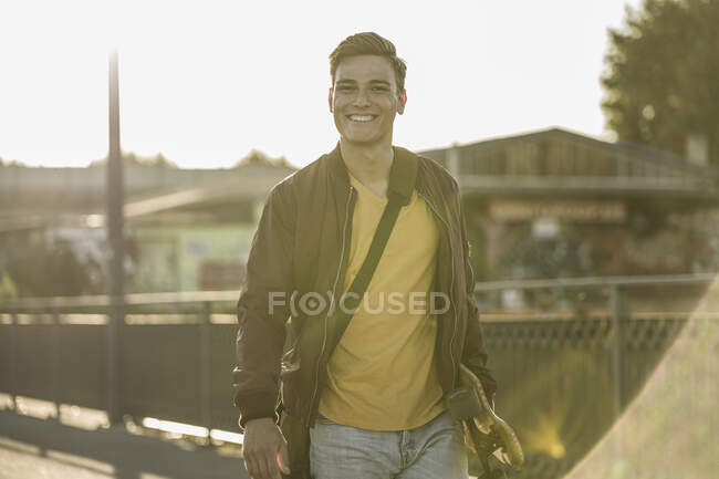 Jovem sorridente carregando skate enquanto estava na cidade durante o dia ensolarado — Fotografia de Stock