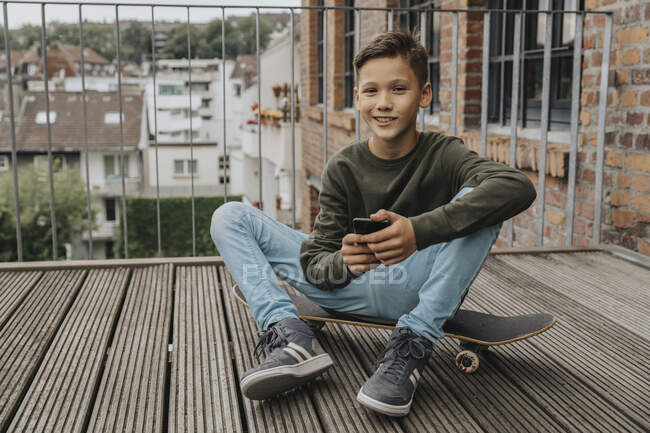 Усміхнений хлопчик використовує смартфон, сидячи на скейтборді проти перил — стокове фото