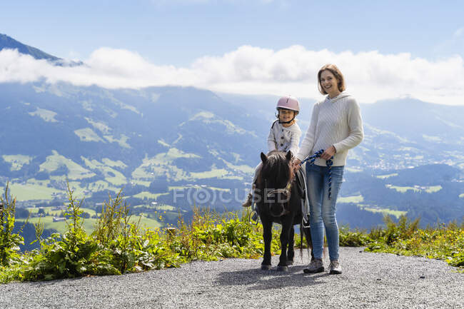 Мать, стоящая рядом с маленькой дочерью верхом на пони против горной долины — стоковое фото