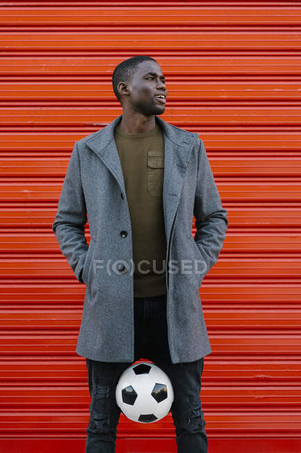 Sorrindo homem segurando bola de futebol entre as pernas enquanto em pé contra a parede vermelha — Fotografia de Stock