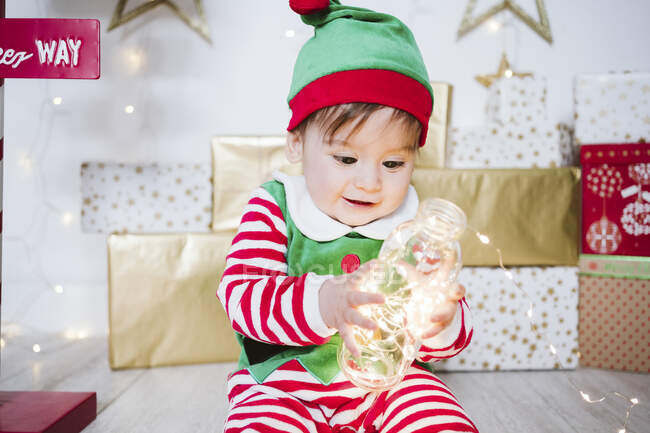 Bambino in costume da elfo che gioca con luci di corda illuminate in bottiglia mentre è seduto a casa durante il Natale — Foto stock