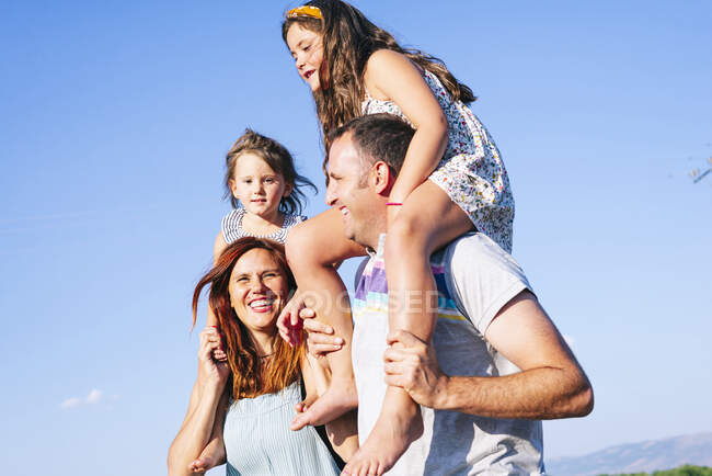Padres alegres llevando hijas en hombros contra el cielo despejado durante el día soleado - foto de stock