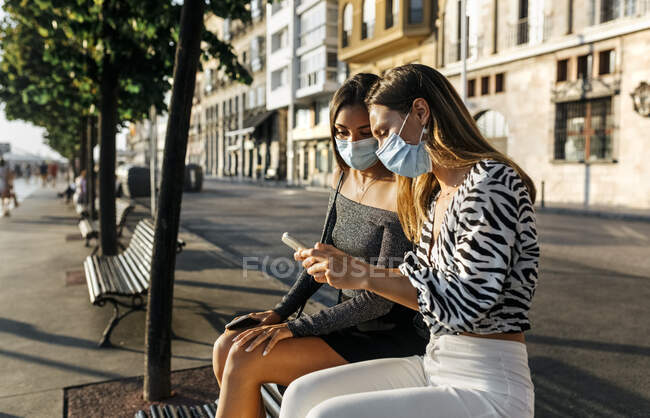 Jóvenes amigos con mascarilla usando el teléfono mientras están sentados en el banco en el paseo marítimo - foto de stock