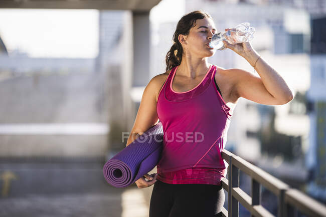 Jovem mulher segurando tapete de exercício beber água enquanto está de pé por trilhos no terraço — Fotografia de Stock