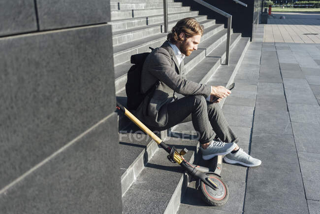 Male commuter using digital tablet while sitting with electric push scooter on steps at downtown — Foto stock