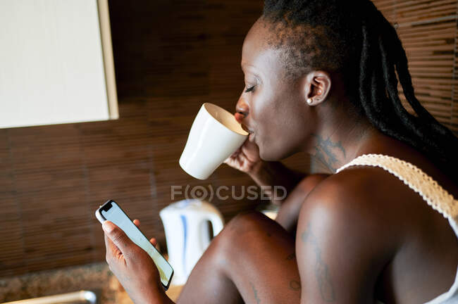 Junge Frau trinkt Kaffee, während sie ihr Smartphone in der Küche benutzt — Stockfoto