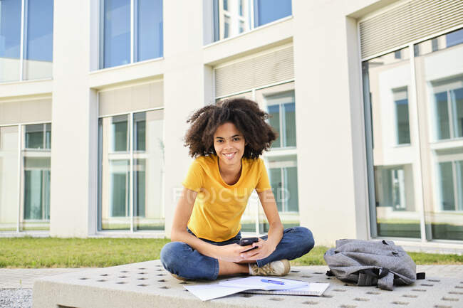 Lächelnder Student mit Büchern per Handy während er auf dem Universitätscampus sitzt — Stockfoto