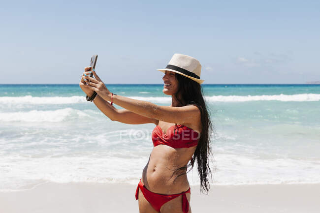 Mujer usando bikini tomando selfie con teléfono inteligente mientras está de pie contra el mar - foto de stock