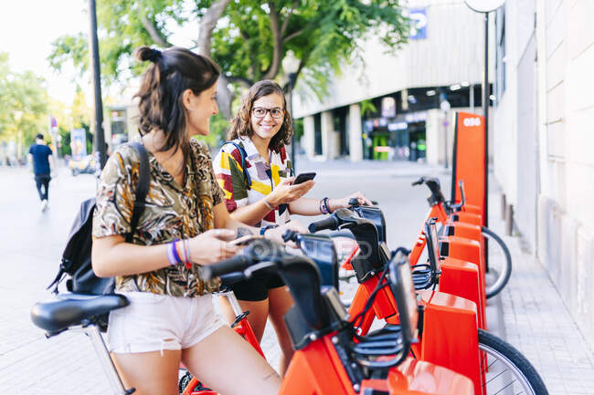 Novias usando teléfono móvil mientras están de pie en la estación de estacionamiento de bicicletas - foto de stock
