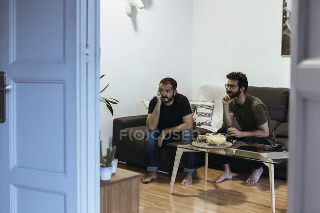 Stressato amici maschi guardando lo sport mentre seduto in soggiorno visto attraverso la porta — Foto stock