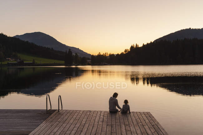 Mutter und kleine Tochter sitzen in der Abenddämmerung gemeinsam am Ufer des Sees — Stockfoto
