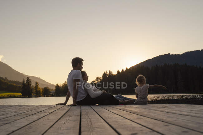 Famille avec petite fille assise ensemble à la fin de jetée au bord du lac au coucher du soleil — Photo de stock