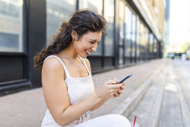 Cheerful young woman using smart phone while sitting on steps in city — Fotografia de Stock