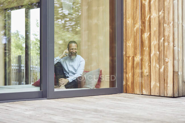 Cheerful man talking over smart phone while sitting on bed in tiny house seen through window — Stock Photo
