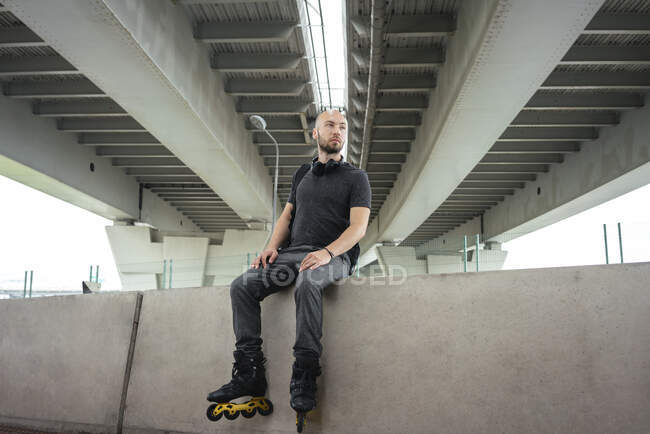 Thoughtful young man wearing inline skates while sitting on retaining wall under bridge - foto de stock