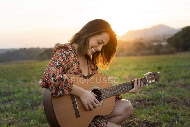 Sorridente giovane donna che suona la chitarra sul prato durante il tramonto — Foto stock