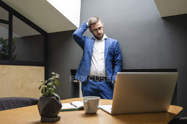 Verwirrter Geschäftsmann kratzt am Kopf, während er auf Laptop auf Schreibtisch im Kreativbüro schaut — Stockfoto