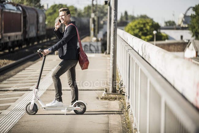 Молодой человек разговаривает по мобильному телефону, стоя со скутером на платформе железнодорожного вокзала — стоковое фото