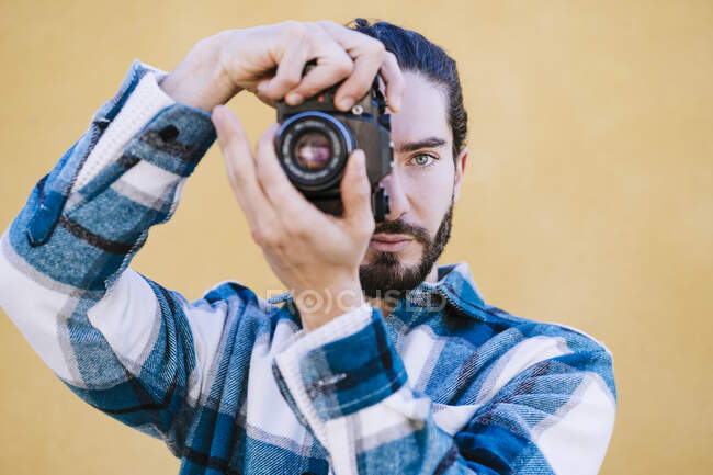 Молодой человек фотографирует через камеру, стоя напротив желтой стены — стоковое фото